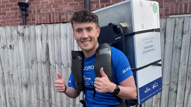 A smiling Jordan Adams runner wearing a blue shirt gives two thumbs up while carrying a large, fridge-like box strapped to his back, standing in front of a wooden fence.