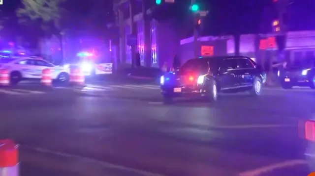 a black limousine rounds a street corner with flashing police lights behind it