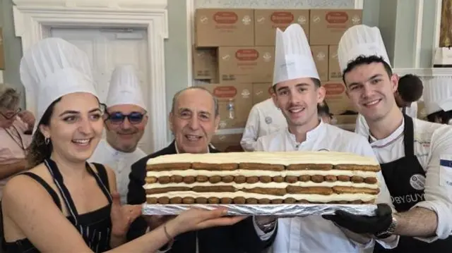A group of five people smile while holding a tiramisu