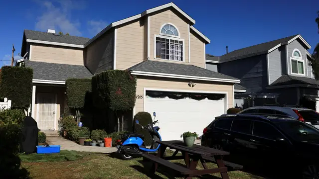 A house with a picnic table and a blue motorbike in front of it. Black and silver cars are parked on the driveway to the right