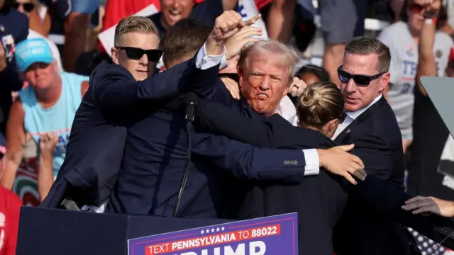 Security officals shield Trump, who raises his fist as a stream of blood streaks down the right side of his face