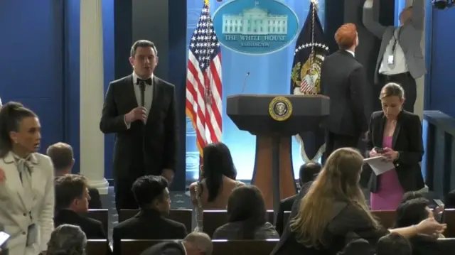reporters sitting in chairs and standing around, some in tuxes and formal attire, in the White House briefing room