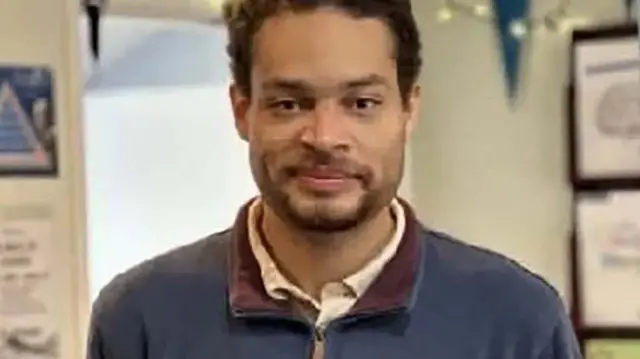 Cole Tomas Allen looks directly at the camera with a slight smile. He is wearing an open-neck shirt and blue jacket.
