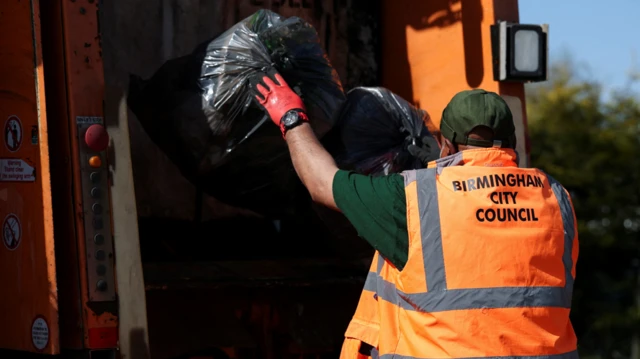 A person with a green t shirt and a green cap with a high vis jacket on is loading bin bags into the back of a bin lorry. You can only see the back of his head and the bin bag which he is throwing into the lorry.