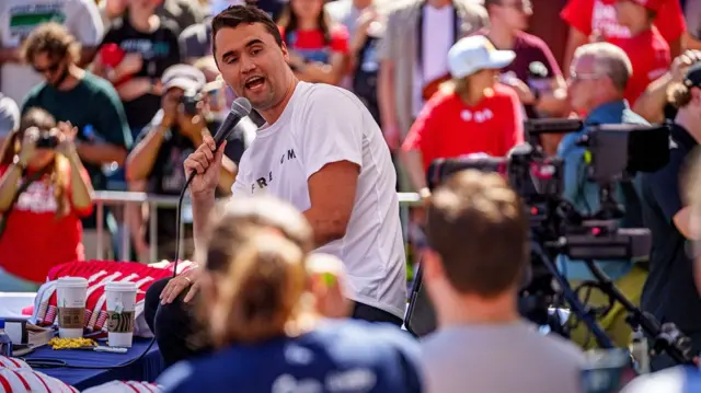 Charlie Kirk wears a white shirt and red hat. He is sitting down and holding a microphone, addressing a crowd of people.
