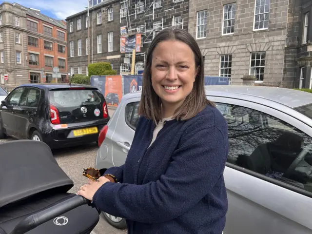 A woman with shoulder length dark hair out for a walk with her baby in a city