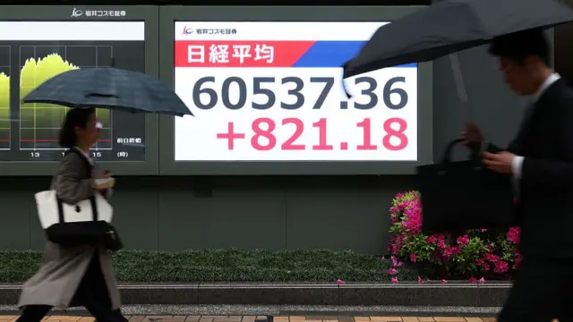 Two people walking past a sign that says 60537.36 and +821.18 in Japan.