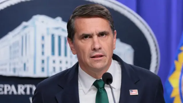 A middle-aged man in a dark suit and green tie stands behind a microphone in front of the seal of the Department of Justice
