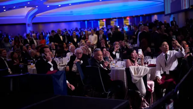 A photo from the White House Correspondents' Dinner, where we can see lots of people stood and gathered around tables. Many have their phones out and are either filming or calling people .
