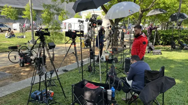 A bank of cameras, tripods and umbrellas stand on an area of grass in the shade. One journalist is sitting on a chair looking at a screen, while another watches over his shoulder.