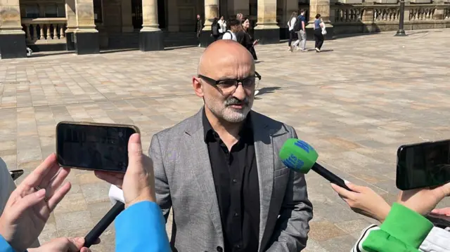 Onay Kasab, a man with a grey beard and bald head, stands in front of phones and microphones