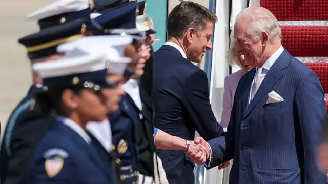 The King shakes the hand of someone waiting in line to greet him. The Queen can just be seen behind him