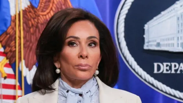 A woman with brown hair and a white suit is shown from the shoulders up in front of flags and an official seal