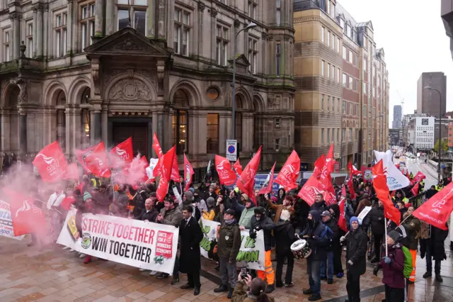 A group of around 100 people with red flags and big white signs are standing on a group. There is also a large amount of smoke in the air. The signs say Unite and Strike Together Win Together.