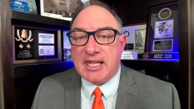 A man speaks to a camera in a virtual interview. He's wearing a suit and tie and behind him are several awards.