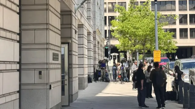 People stand outside of a US Courthouse. Some have camera on tripods.
