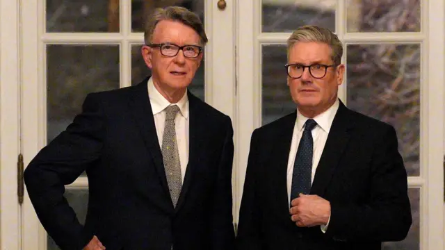 Mandelson (L) and Starmer (R) stand together at the bottom of a corridor. Both are dressed in suits and black-rimmed glasses
