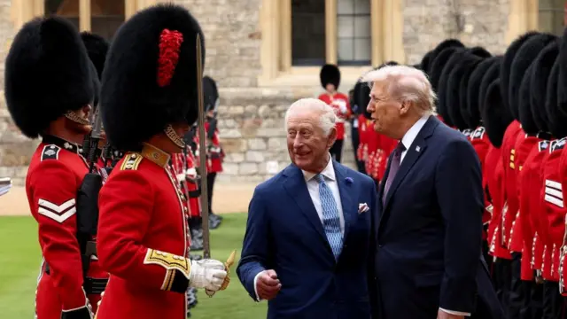 King Charles and Donald Trump stood next to rows of beefeater guards wearing red jackets and large black hats. They're talking to one of the guards.