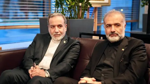 Iranian foreign minister Abbas Araghchi in a suit sat on sofa next to a man in a black suit.