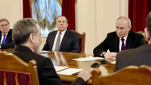 President Putin sat behind a table, facing Abbas Araghchi. Both are wearing dark suits, Araghchi's back is to the camera. Two other men are sat next to Putin.