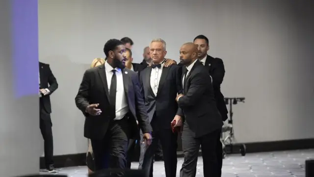 Health and Human Secretary Robert Kennedy Jr. is evacuated by four law enforcement officers on each corner of him while his wife follows beside, next to one of the enforcement officers
