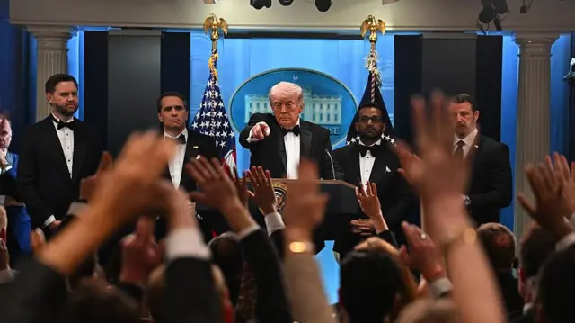 Trump, wearing a bow tie and tuxedo, points to a reporter in a room full of raised hands trying to get his attention.