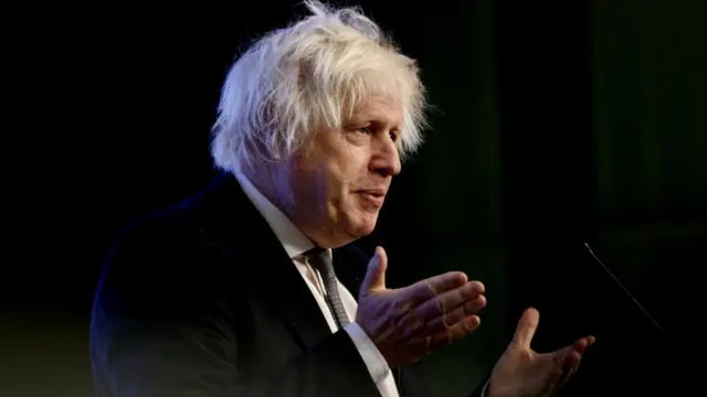 Boris Johnson speaks at a microphone while wearing a dark suit and grey tie