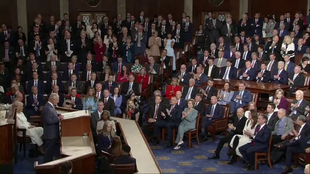 A view of the King speaking with dozens of audiences members in the background.