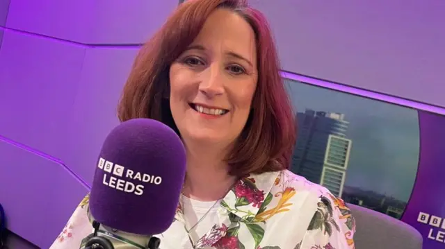 A woman with red-brown hair smiling and sitting in front of a purple microphone which says BBC Radio Leeds