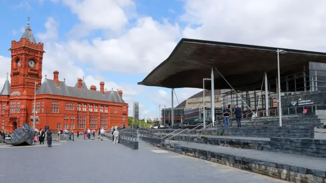 A view of the Senedd, the Welsh parliament building in Cardiff with the Pierhead Building in the background. Members of the public are milling around in front of the building