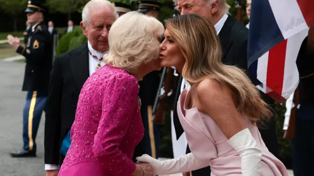 Queen Camilla and Melania Trump kiss on the cheek in greeting, wearing complementary shades of pink.