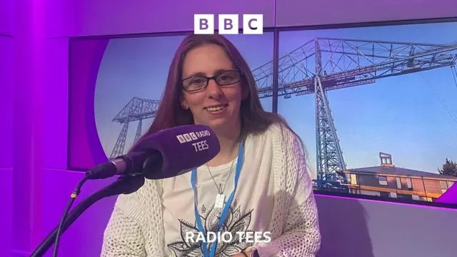 A woman with long brown hair and glasses smiles at the camera, she wears a white top with a black and white flower on and sits in a BBC Radio Tees purple studio. The microphone in front of her says 'BBC Radio Tees' and the backdrop shows a bridge.