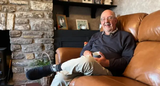 John Sibley is smiling at the camera and has one leg crossed over the other while sitting on a brown leather sofa. He has grey hair and a beard and is wearing a navy sweatshirt and cream chinos.