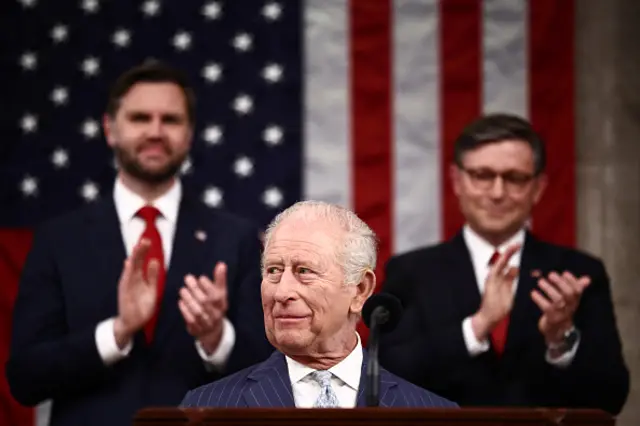 King Charles stands at a podium with JD Vance and Mike Johnson clapping behind him.
