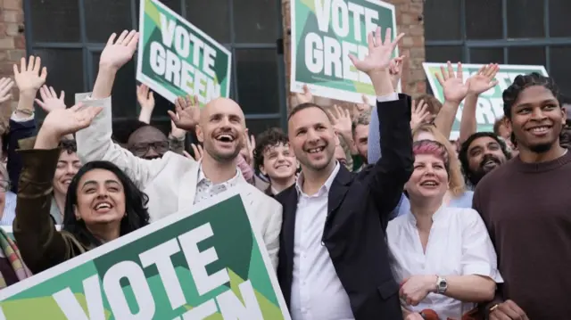 A crowd of people - including men and women - with Zack Polanksi in the centre are looking out to the distance smiling and waving. Some are holding large "VOTE GREEN" signs
