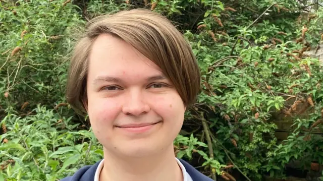 A young white man (John Blandamour) with brown hair, smiles for the camera against a leafy backdrop