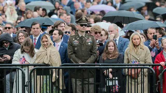 Several people stand at a barrier in the rain. The figure in the middle appears to be wearing a formal US Army uniform. Many are holding black umbrellas