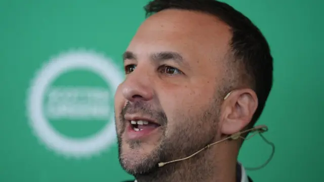 A portrait photo of a man with a head mic who is slightly smiling. He has dark, short hair and a light beard. Behind him is a green background with a logo which is blurred out