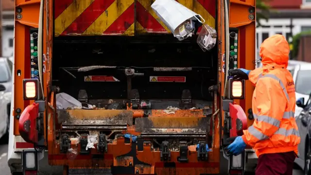 Waste is collected by refuse worker in Aston, Birmingham. They are wearing an orange hi-vis jacket and blue gloves as they throw rubbish into the back of a bin wagon