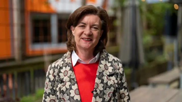 Eluned Morgan smiles at the camera. She has mid length brown hair and weares a flowery coat and red jumper.