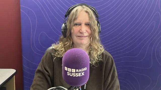 Sarah Gorell, who has long wavy hair and is wearing a dark top and headphones, sits behind a purple BBC Radio Sussex microphone. There is a purple backdrop behind her
