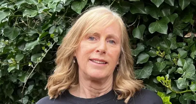 A woman looks into the camera smiling. She has long, blonde hair and blue eyes, and is wearing a dark blue top. Behind her are dark green ivy leaves