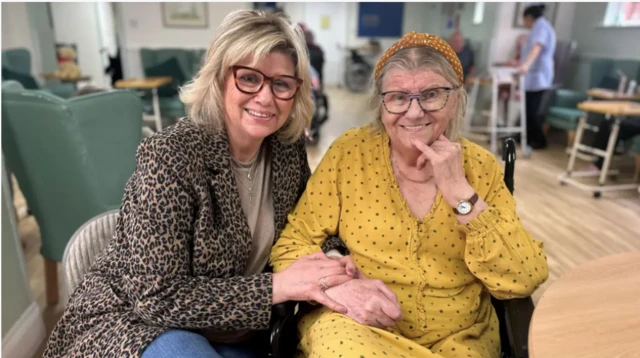 Leonie Williams has blonde shoulder-length hair and is wearing a taupe top and leopard print jacket and is holding onto her mother. Angela Martens is wearing a yellow and black spotted dress and a yellow headband. They are sitting down.