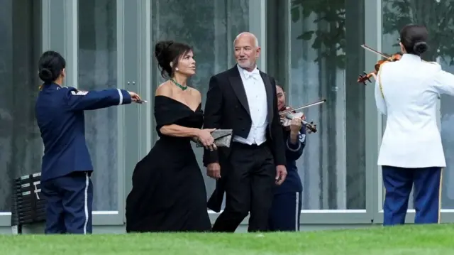 Jeff Bezos in white tie and tuxedo, accompanied by his wife Lauren Sanchez in a formal black gown, walk between violinists outside the White House