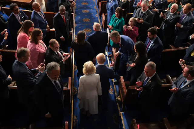 The King enters the chamber with Queen Camilla and people clapping either side.