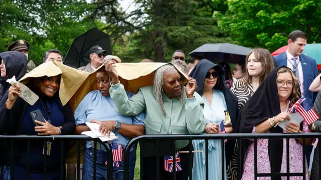 People stand at a barricade while sheltering from rain. The woman in the middle is wearing a light green jacket and is holding a tarpaulin over herself and two others. Several individuals are holding union jack flags, as well as USA flags.