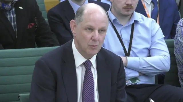 Barton giving evidence to committee. He is wearing a suit and purple tie