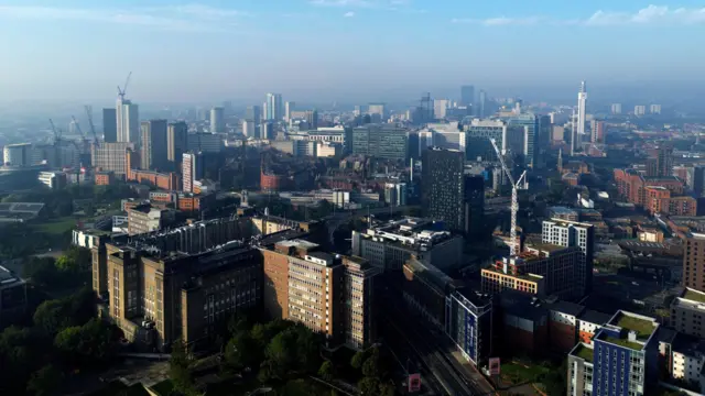 An aerial view of Birmingham city centre. A variety of high-rise buildings, cranes and a slight mist.