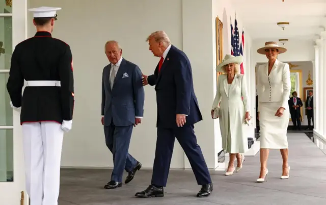 A marine stands guard with his back to the camera as two men in suits walk by with two women in ligth coloured formal wear and hats follow