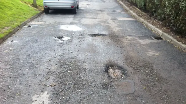 Potholes on a small road, with the back of a car in the background